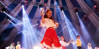 menina no palco em destaque no meio da foto dançando feliz com outras em volta com vestidos iguais; outros rapazes dançando junto e feixes de luzes azul