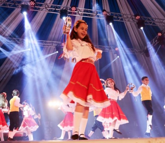menina no palco em destaque no meio da foto dançando feliz com outras em volta com vestidos iguais; outros rapazes dançando junto e feixes de luzes azul