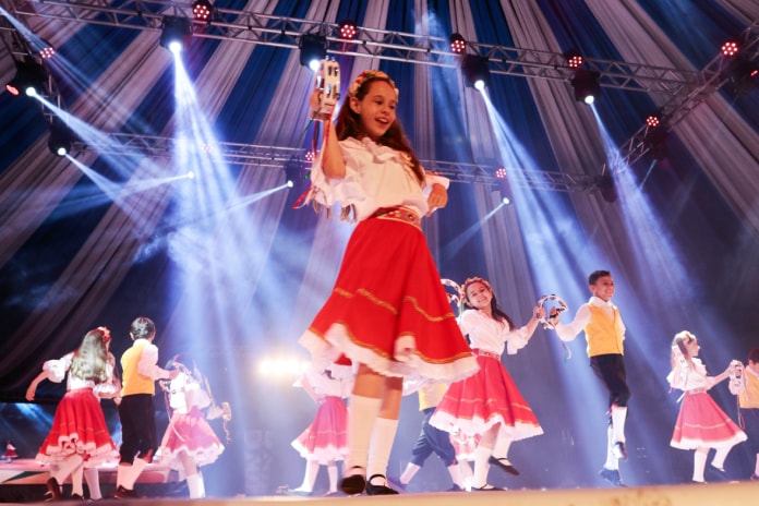 menina no palco em destaque no meio da foto dançando feliz com outras em volta com vestidos iguais; outros rapazes dançando junto e feixes de luzes azul
