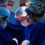6 médicos vestindo jaleco azul, com toucas na cabeça, realizando uma cirurgia - mutirão para atender demanda de cirurgias eletivas em sc