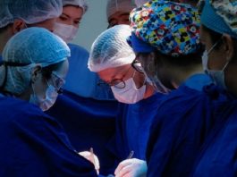 6 médicos vestindo jaleco azul, com toucas na cabeça, realizando uma cirurgia - mutirão para atender demanda de cirurgias eletivas em sc