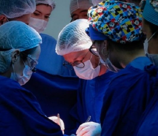 6 médicos vestindo jaleco azul, com toucas na cabeça, realizando uma cirurgia - mutirão para atender demanda de cirurgias eletivas em sc