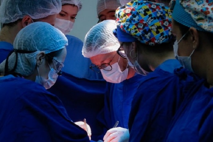 6 médicos vestindo jaleco azul, com toucas na cabeça, realizando uma cirurgia - mutirão para atender demanda de cirurgias eletivas em sc