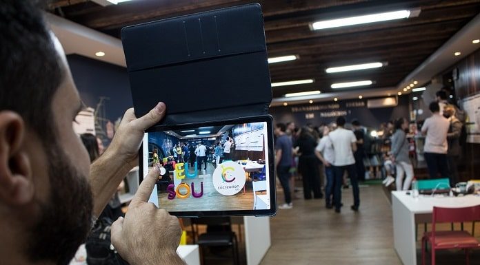 homem bate foto com tablet do ambiente do cocreation lab, onde há mesas e pessoas em pé no fundo