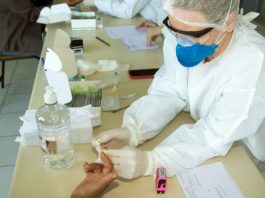 profissional de saúde faz teste no dedo de pessoa com mão apoiada sobre mesa onde estão outros objetos