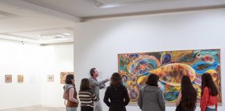 seis mulheres em pé, de costas para a foto, olham um homem falando e apontando para quadro em exposição no masc