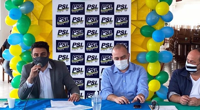 fernando anselmo sentado atrás de bancada com bandeira do brasil fala ao microfone; ele usa terno e máscara; ao lado sentados outros dois homens de máscara; ao fundo painel do partido com balões verdes, amarelos e azuis