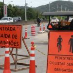 placas de sinalização de obra às margens da rodovia, indiciando entrada e saída de caminhões e passagem de pedestres e cilclistas; rodovias e carros ao fundo