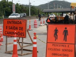 placas de sinalização de obra às margens da rodovia, indiciando entrada e saída de caminhões e passagem de pedestres e cilclistas; rodovias e carros ao fundo