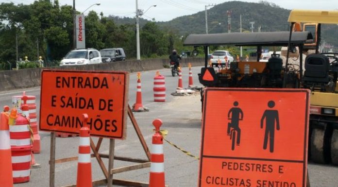 placas de sinalização de obra às margens da rodovia, indiciando entrada e saída de caminhões e passagem de pedestres e cilclistas; rodovias e carros ao fundo