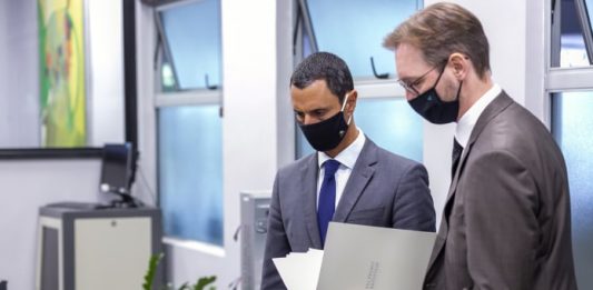 Moisés e Daniela entregam à Alesc defesas contra impeachment amin e probst ambos usando terno e máscara olham para baixo na mesma direção; probst com documento aberto na mão; estão em uma sala de escritório