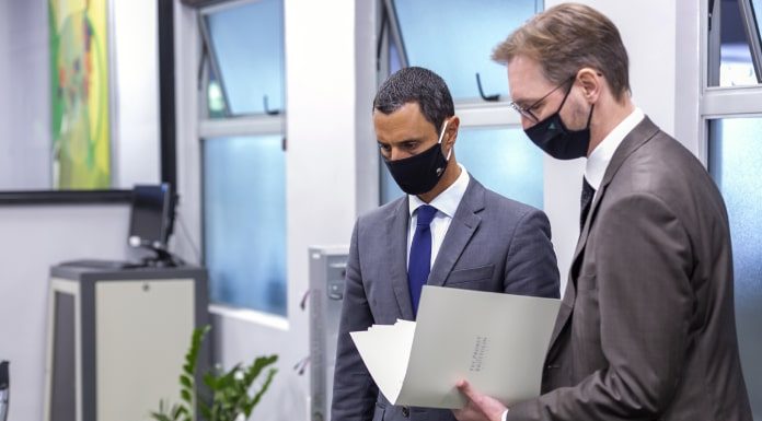 amin e probst ambos usando terno e máscara olham para baixo na mesma direção; probst com documento aberto na mão; estão em uma sala de escritório