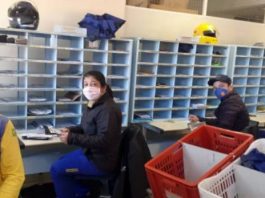 quatro funcionários dos correios usando máscaras se viram para olhar para a foto; eles estão sentados em frente a diversos nichos de cartas