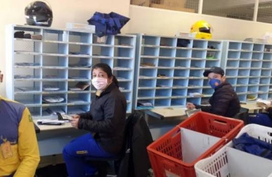 Funcionários dos Correios em SC aceitam parcialmente decisão do TST e finalizam greve quatro funcionários dos correios usando máscaras se viram para olhar para a foto; eles estão sentados em frente a diversos nichos de cartas