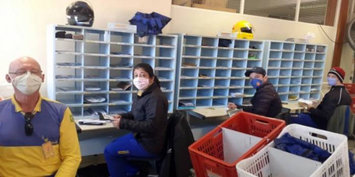 quatro funcionários dos correios usando máscaras se viram para olhar para a foto; eles estão sentados em frente a diversos nichos de cartas