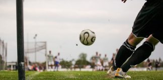 jogador inclinado logo após chutar a bola de escanteio, que viaja em direção à área do gol ao fundo, onde estão outros jogadores