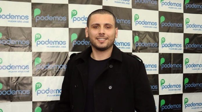 hélio costa jr olha com sorriso para a câmera usando blazer preto e em frente a um painel com logos do podemos