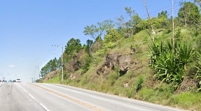 terreno íngreme com rochas e vegetação na margem da rodovia; carro ao fundo em cima do morro