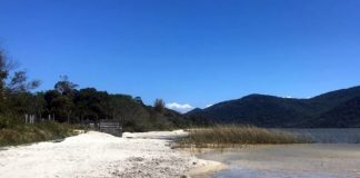 praia de areia na margem da lagoa com vegetação ao centro, ao lado e montanhas ao fundo na outra margem