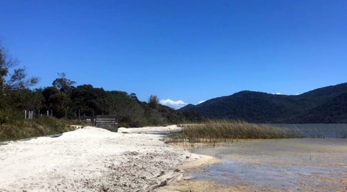 praia de areia na margem da lagoa com vegetação ao centro, ao lado e montanhas ao fundo na outra margem