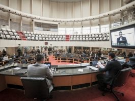 deputados vistos de trás sentados na bancada da mesa diretora da alesc com o plenário circular vazia ao fundo; amin em transmissão no telão ao lado