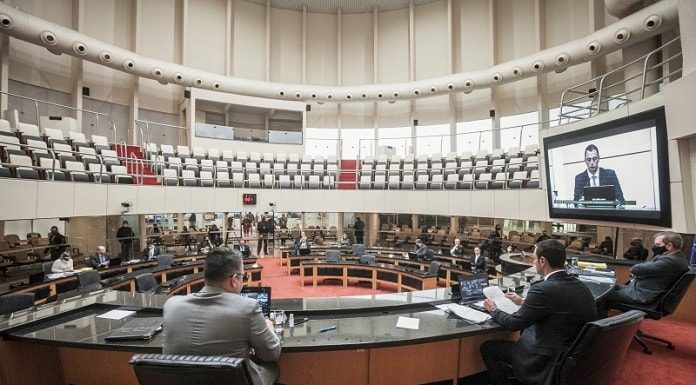 deputados vistos de trás sentados na bancada da mesa diretora da alesc com o plenário circular vazia ao fundo; amin em transmissão no telão ao lado