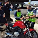 policial e funcionários da als ao redor de moto instalando antena corta pipa e outro homem de máscara em pé ao lado; carros ao fundo