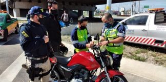 policial e funcionários da als ao redor de moto instalando antena corta pipa e outro homem de máscara em pé ao lado; carros ao fundo