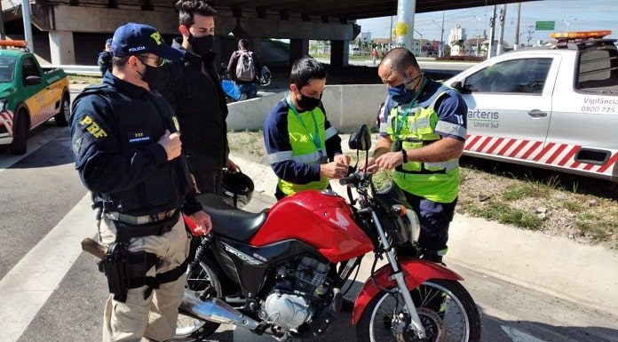 policial e funcionários da als ao redor de moto instalando antena corta pipa e outro homem de máscara em pé ao lado; carros ao fundo