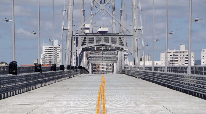 pista da ponte hercílio luz vista de frente com linha divisória de tráfego ao longo da estrutura; base da torre no fundo da foto; prédios ao fundo