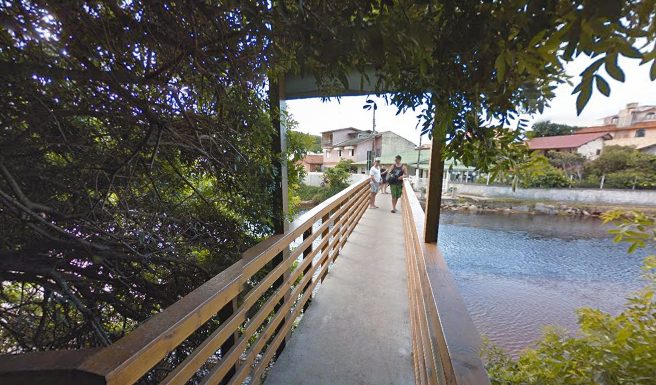 ponte do rio sangradouro vista da margem com guarda-corpo alto de madeira, pequeno portal de madeira e árvores sobre a cabeceira; casal no meio da ponte