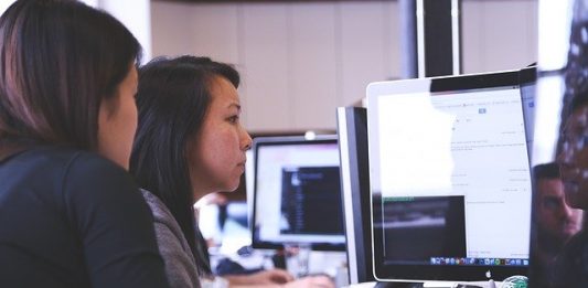 Vagas no setor de tecnologia catarinense Duas mulheres sentadas a esquerda, de frente para um computador a direita