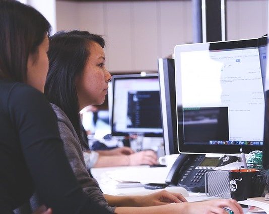Duas mulheres sentadas a esquerda, de frente para um computador a direita