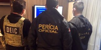 três agentes vistos de costas em pé em sala de apartamento olhando par uma televisão; as inscrições nos casacos de cada um são Dei, Perícia Oficial e Oficial IGP