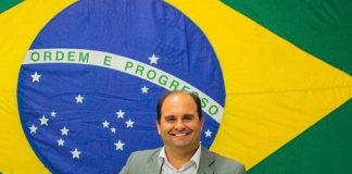 sergui guimarães olha sorridente para a foto; atrás há uma grande bandiera do brasil; ele usa camiseta e blazer cinza