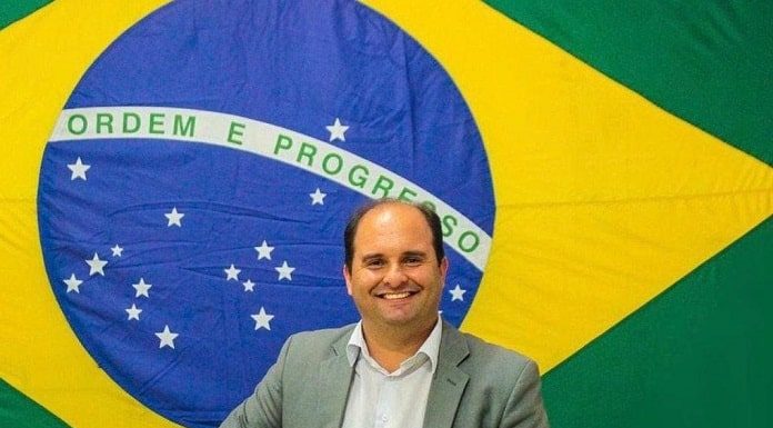 sergui guimarães olha sorridente para a foto; atrás há uma grande bandiera do brasil; ele usa camiseta e blazer cinza