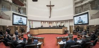 plenário visto pelo meio da bancada com grande cruz no alto e deputados olhando presidente do tjsc no telão