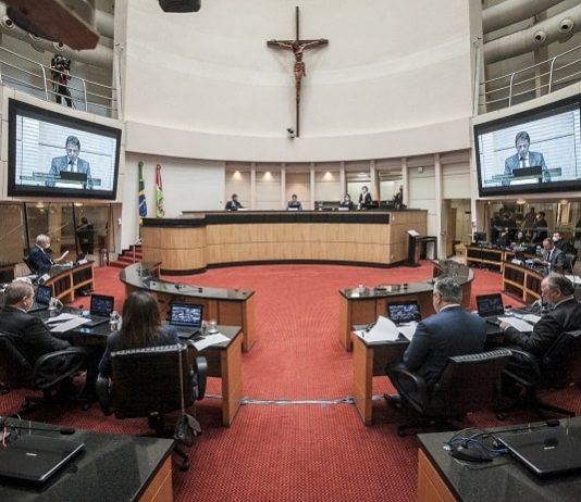 plenário visto pelo meio da bancada com grande cruz no alto e deputados olhando presidente do tjsc no telão