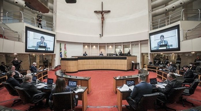 plenário visto pelo meio da bancada com grande cruz no alto e deputados olhando presidente do tjsc no telão