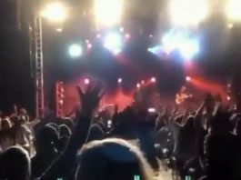 público visto de costas em show, aparentemente pessoas sentadas; palco ao fundo