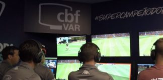 homens vistos de costas sentados à frente de monitores com imagens de jogo e inscrição "CBF VAR" em suas costas