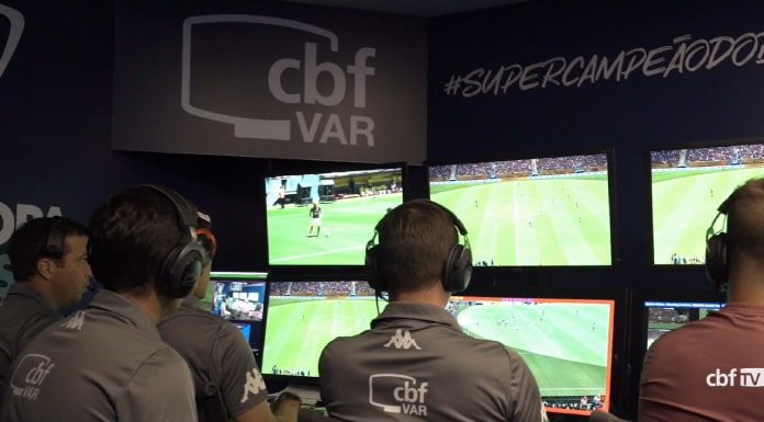 homens vistos de costas sentados à frente de monitores com imagens de jogo e inscrição "CBF VAR" em suas costas