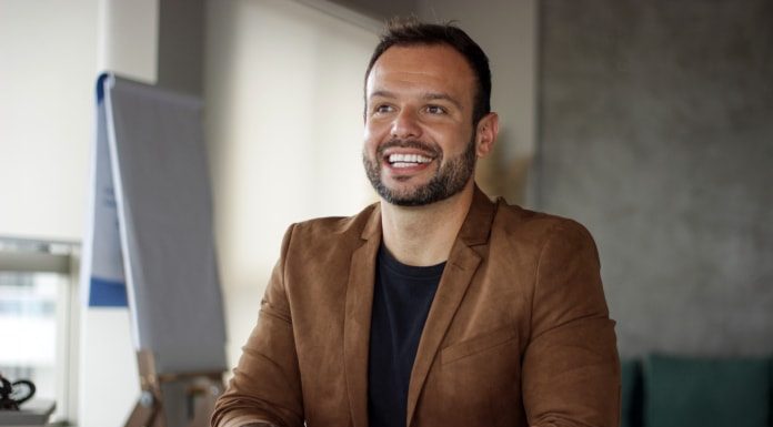 pedrão sorridente olha um pouco para o lado; ele está sentado em uma mesa de escritório, usando um blazer marrom