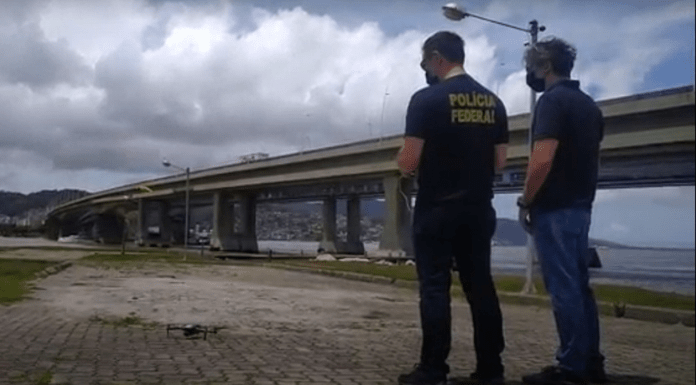 dois policiais federais de costas olham para drone no céu ao lado das pontes de florianópolis