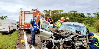 carro completamente destruído de frente; bombeiros e socorristas em volta; caminhão de bombeiros ao fundo