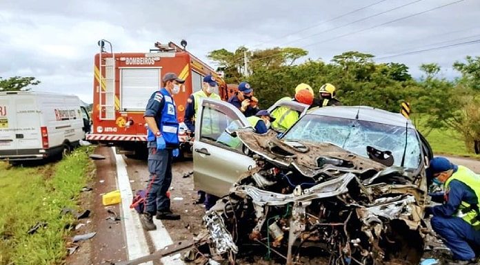carro completamente destruído de frente; bombeiros e socorristas em volta; caminhão de bombeiros ao fundo