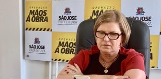 adeliana sentada com braços cruzados sobre mesa onde olha para um celular à sua frente em pé; painel de logos da prefeitura ao fundo