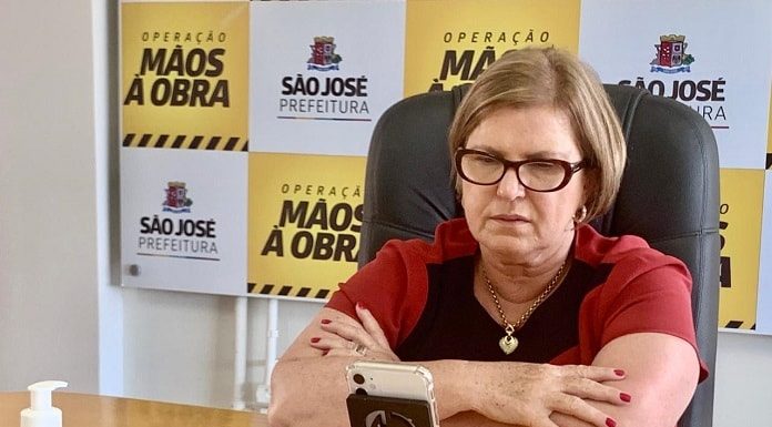 adeliana sentada com braços cruzados sobre mesa onde olha para um celular à sua frente em pé; painel de logos da prefeitura ao fundo