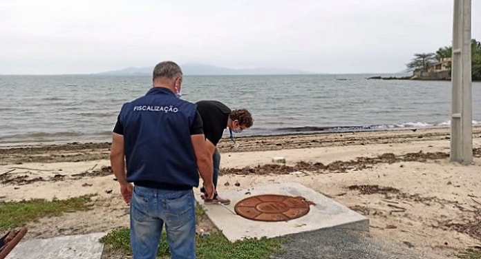 dois fiscais usando coletes próximos a tampa de bueiro na areia de uma praia, em dia nublado e mar encrespado