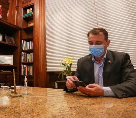 moisés usa máscara e mexe em celular sobre mesa de mármore; livros em estante ao lado